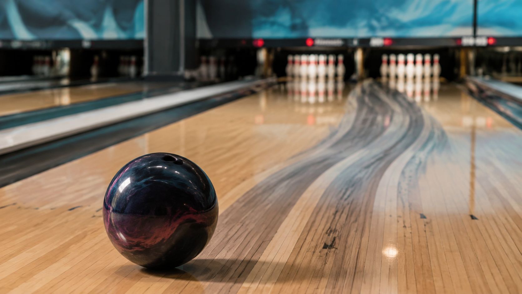A bowling ball resting on a polished wooden bowling lane in front of a full set of pins.