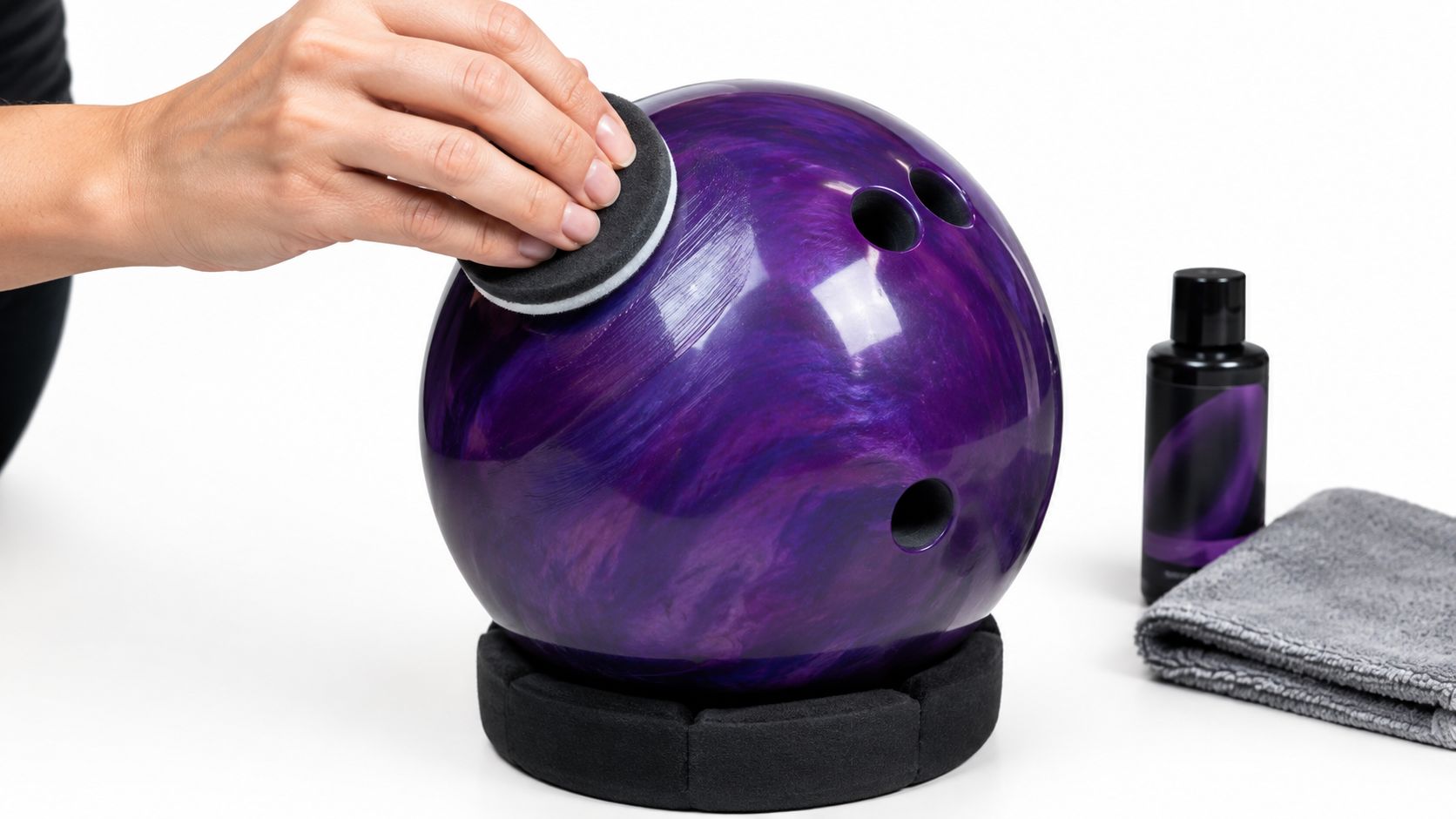 A hand using a foam pad to polish a purple marbled bowling ball on a stand.